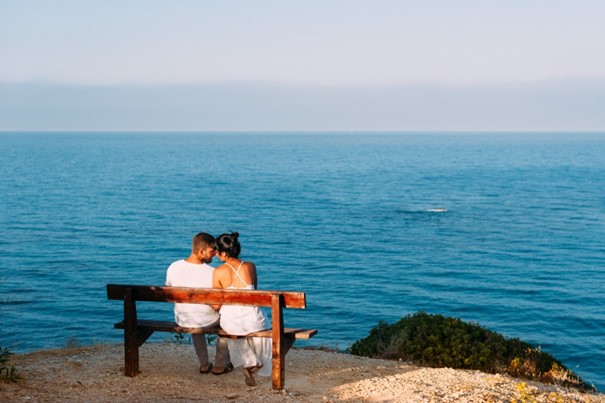 the back of a couple sitting on bench in front of ocean